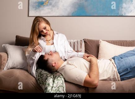 Zärtlichkeit, Liebeskonzept. Paar entspannen sich auf der Couch und schauen sich gegenseitig an. Hübscher Mann, der auf dem Sofa liegt und den Kopf auf die Knie einer schönen Frau setzt. Heimatdatum. Hochwertige Fotos Stockfoto