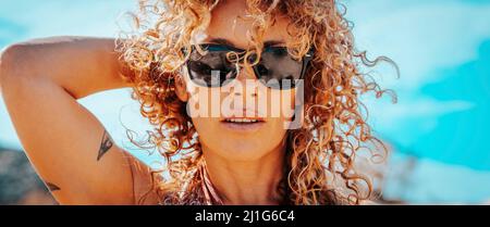 Sommerportrait einer schönen jungen erwachsenen Frau mit Sonnenbrille und gesundem blonden langen lockigen Haaren, die in die Kamera schaut. Attraktive Brünette mit blauem Himmel Stockfoto