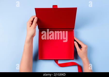 Weibliche Hände öffnen leere rote elegante Luxus-Box mit Band. Mockup Geschenk Geschenkbox auf hellblauem Hintergrund. Draufsicht Stockfoto