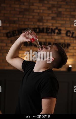 RIVNE, UKRAINE JUNI 26 2020: Junger bärtiger Mann trinkt Coca Cola aus der Flasche im Friseurladen auf Backsteinmauer Hintergrund. Stockfoto