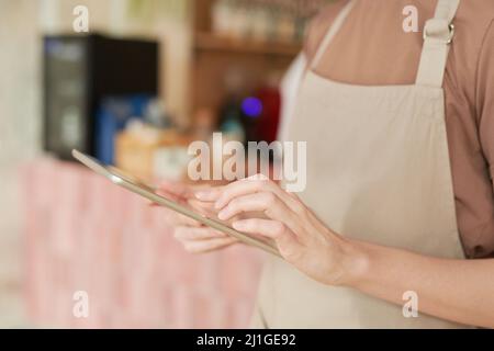 Horizontale Nahaufnahme mit mittlerem Ausschnitt einer nicht erkennbaren Frau mit Schürze, die mit einem digitalen Tablet im Café arbeitet Stockfoto