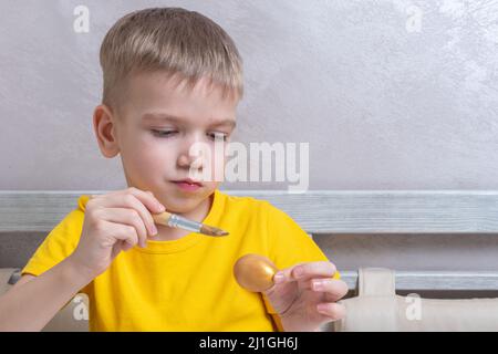 Ein kleiner blonder Junge malt Eier für die Osterferien zu Hause. Das Kind hat Spaß und feiert den Urlaub. DIY Ostereier Konzept. Stockfoto