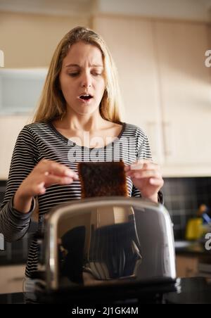 Es sieht so aus, als würde es einer dieser Tage sein. Aufnahme einer jungen Frau, die zu Hause eine Scheibe verbranntem Toast aus einem Toaster entfernt hat. Stockfoto