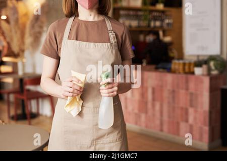 Horizontaler mittlerer Ausschnitt einer nicht erkennbaren jungen Kellnerin, die eine Maske mit Desinfektionsspray und Wischtuch trägt Stockfoto