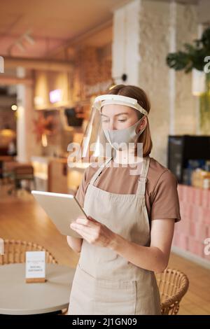 Mittellange Porträtaufnahme einer jungen Kellnerin mit Schürze und Schutzmasken auf dem Gesicht, die mithilfe eines digitalen Tablets Bestellungen online überprüft Stockfoto