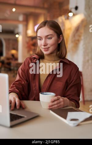Vertikale mittlere Porträtaufnahme einer stilvollen kaukasischen Frau, die während der Kaffeepause am Kaffeetisch sitzt und einen Videoanruf auf dem Laptop führt Stockfoto