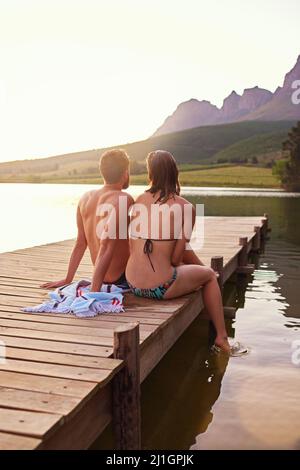Ruhe und Frieden. Rückansicht eines liebevollen jungen Paares in Badeanzügen, das bei Sonnenuntergang auf einem Dock sitzt. Stockfoto