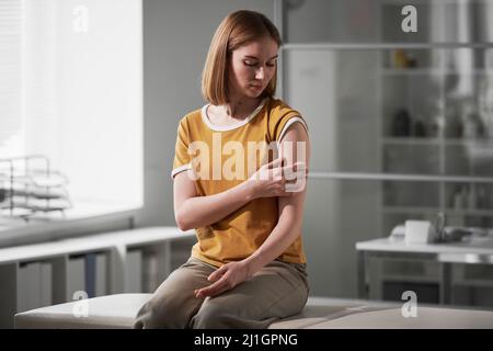 Horizontales mittellange Porträt einer jungen kaukasischen Frau, die im Impfraum sitzt und das Wattepad auf die Schulter drückt Stockfoto
