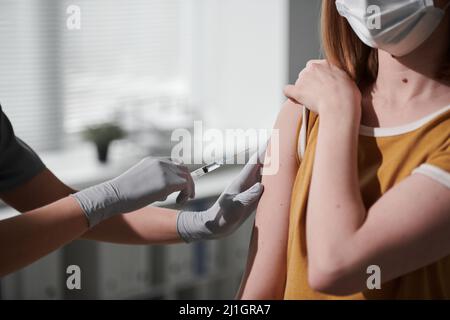 Mittlere Nahaufnahme eines nicht erkennbaren Arztes mit Latexhandschuhen, der die Spritze hält und eine weibliche Patientin impft Stockfoto
