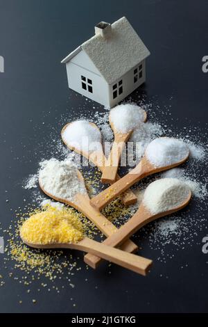 Teelöffel aus Holz mit Mehl, Salz, Zucker, Grieß und Maismehl, Führt zu einem kleinen weißen Haus Stockfoto