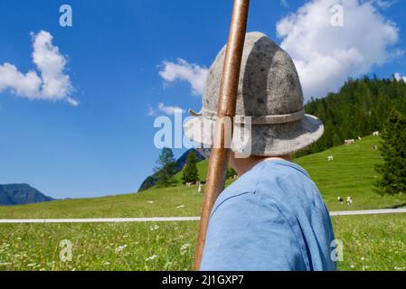 Kleiner Junge mit traditionellem Hut und Holzstab hilft, Kühe in den österreichischen Alpen zu hüten. Vorarlberg, Österreich. Stockfoto