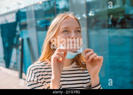 Die europäische glückliche 30s-Frau zieht ihre Maske am sonnigen Frühlingstag auf der Straße aus Stockfoto