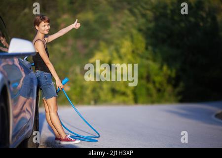 Mädchen, das neben einem Elektroauto steht und ein Ladekabel hält und mit dem Anhalter unterwegs ist. Stockfoto