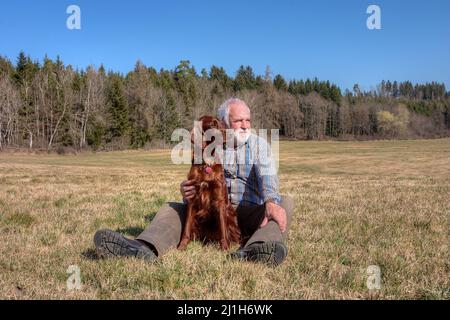 Ein Mann sitzt auf einer Wiese. Sein irischer Setter-Hund sitzt zwischen seinen Beinen. Stockfoto