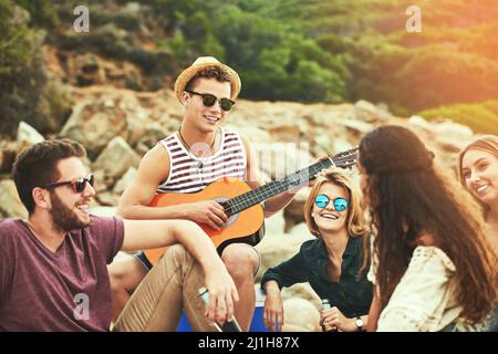 Es ist immer ein Vergnügen, für euch zu spielen. Aufnahme eines jungen Mannes, der für seine Freunde Gitarre spielt, während er am Strand chillte. Stockfoto