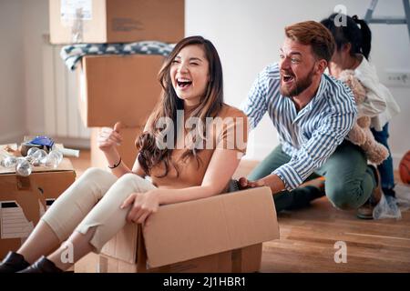 Schöne junge asiatische junge Frau sitzt in Karton, lacht, spielt mit ihrem Mann und ihrer Tochter in einer neuen Wohnung. Junge Familie Konzept Stockfoto