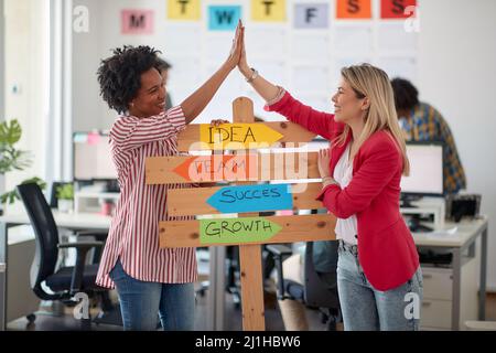 Zwei junge, fröhliche Büroangestellte haben eine gute Zeit, während sie für ein Foto posieren und Firmenslogans in einer angenehmen Atmosphäre in der fördern Stockfoto