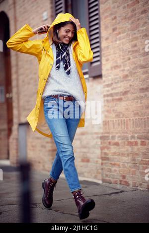 Eine junge, hübsche Frau im gelben Regenmantel ist gut gelaunt, wenn sie an einem regnerischen Tag entspannt durch die Stadt geht. Spaziergang, Regen, Stadt Stockfoto