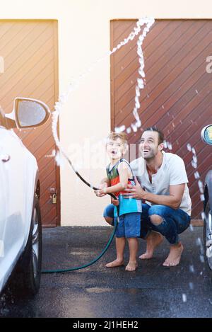 Sie haben so viel Spaß. Die ganze Aufnahme eines Vaters und eines Sohnes, die mit einer Schlauchleitung spielen, während sie ein Auto gemeinsam waschen. Stockfoto