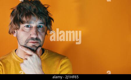 Junger hispanischer Mann in legerer Kleidung, der über Zweifel dachte, mit den Fingern auf dem Kinn, der die Kamera betrat und sich über einen orangenen Rücken wunderte Stockfoto