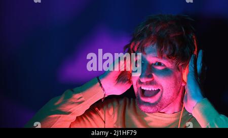 Junger Mann tragen gelbe Hemd Kopfhörer hören Musik singen Song aufnehmen Stimme isoliert auf Neonlicht Farbe Hintergrund Studio Porträt. Stockfoto