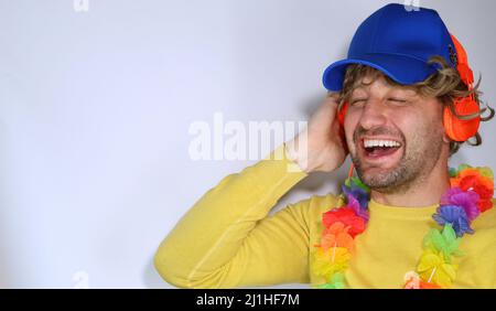 Bild eines gutaussehenden Mannes im lässigen und Partyoutfit, trägt eine blaue Kappe, hört Musik in orangefarbenen Kopfhörern, lächelt zufrieden, glücklich über Weiß stehend Stockfoto