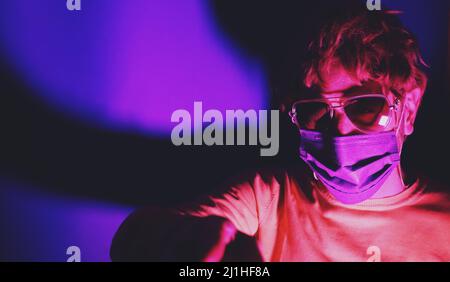Mann mit Sonnenbrille in violetter Gesichtsmaske in Neonlicht auf violettem Betongrund. Lockdown-Design. Stilvolles Outfit während der Quarantäne von Coronaviru Stockfoto
