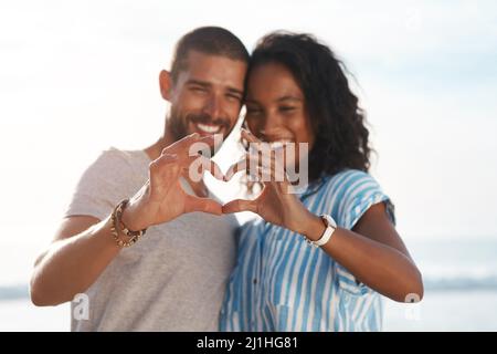 Dein Herz wird nahe an meinem gehalten. Aufnahme eines jungen Paares, das mit den Händen am Strand eine Herzform formt. Stockfoto