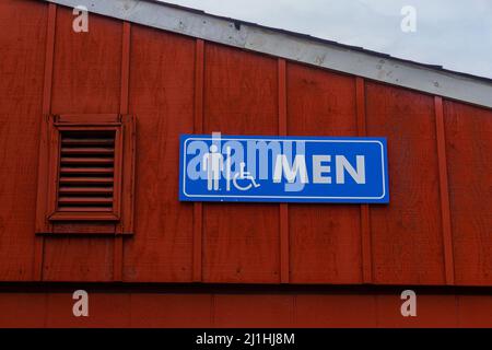 Badschild für Männer auf der Oberseite eines Gebäudes neben dem Dach, blau und weiß vor einer roten Wand. Stockfoto