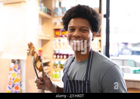 Porträt eines lächelnden jungen afroamerikanischen männlichen Barista, der Brot mit einer servierten Zange im Café hält. Unveränderte Café-Kultur, Menschen und Beruf conce Stockfoto