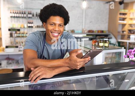 Porträt des lächelnden jungen männlichen afroamerikanischen Besitzer hält Tablet-pc auf Schrank im Café gelehnt. Unverändert, Café-Kultur, Menschen und Beruf con Stockfoto