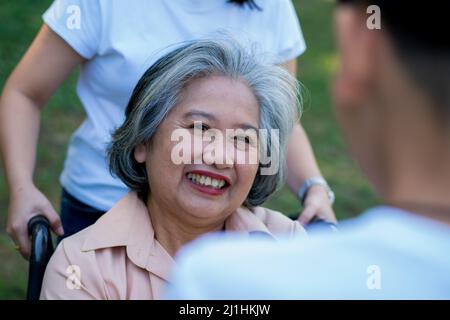 Glückliche ältere asiatische Großmutter benutzt Rollstuhl mit ihrer Tochter und Enkelkind im Park, Enkel kam, um ältere Großmutter zu besuchen und Hand zu halten. Kontra Stockfoto