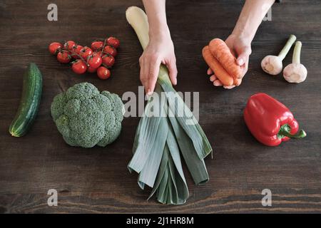 Aus der Sicht der weiblichen Hände, die Lauch-Zwiebeln und Karotten mit anderem Gemüse auf den Holztisch legen Stockfoto