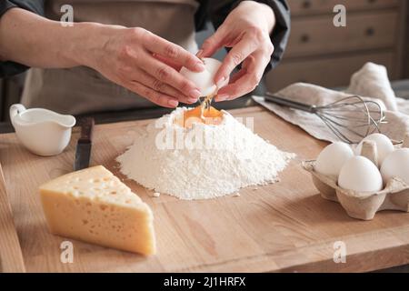 Nahaufnahme einer nicht erkennbaren Frau, die am Tisch steht und das Ei auf einem Holzbrett mit Käse und Schneebesen in Mehl zerbricht Stockfoto