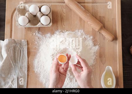 Oberhalb der Ansicht einer unkenntlichen Frau, die Eigelb und Eiweiß mit Schale über Holzbrett mit Mehl trennte Stockfoto