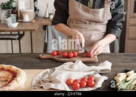 Nahaufnahme einer nicht erkennbaren Frau in der Schürze, die an der Theke mit Gemüse steht und Tomaten auf Holzbrett schneidet Stockfoto