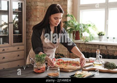 Attraktive Brünette Frau in Schürze, die an der Küchentheke steht und Pizza an Bord anpasst, während sie für das Abendessen zubereitet Stockfoto