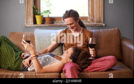 Heres zu uns lieben. Aufnahme eines jungen, entsetzten Paares, das auf dem Sofa Wein trinkt. Stockfoto