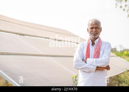 Handgeführte Aufnahme eines lächelnden Dorfbauern, der mit gekreuzten Armen steht, indem er die Kamera vor dem Solarpanel anschaut - Konzept der modernen Landwirtschaft, grün Stockfoto