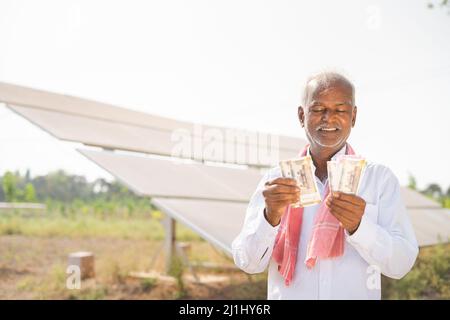 Glücklich lächelnder indischer Landwirt, der Geld couting, während er vor dem Solarpanel auf Ackerland steht - Konzept der Stromeinsparung, Investitionen, Banken Stockfoto