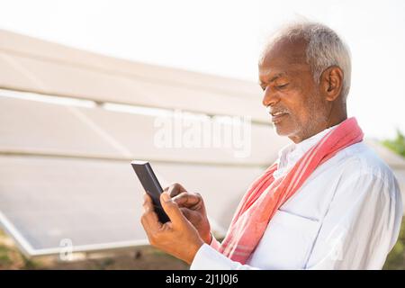 Indischer Landwirt beschäftigt mit dem Handy vor dem Solarpanel auf dem Agrarfeld - Konzept der Technologie, erneuerbare Energien und moderne Landwirtschaft. Stockfoto