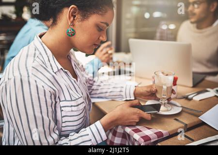 Immer beschäftigt wie üblich. Aufnahme einer fokussierten jungen Frau, die auf ihr Handy schreibt, während sie an einem Coffeeshop sitzt und ein heißes Getränk trinkt. Stockfoto