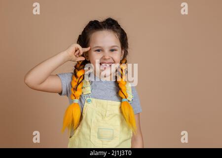 Kleines leuchtendes Kind mit gelben Kanekalon-Zöpfen, mit dem Finger, der vom Tempel gesetzt wird, lächelnd mit fehlenden Zähnen im gelben Jumpsuit und grauem T-Shirt an Stockfoto