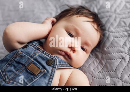 Portrait eines hübschen Babys, das in Nahaufnahme auf einer grauen Decke in Denim-Strampler ruht. Ruhiges Kleinkind lag auf dem Rücken im Schlafzimmer, erkunden Sie die Welt. Grau Stockfoto