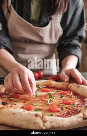 Nahaufnahme einer nicht erkennbaren Frau in der Schürze, die Erbsen auf Pizza mit Tomaten legt, während sie sie für die Familie zubereitet Stockfoto