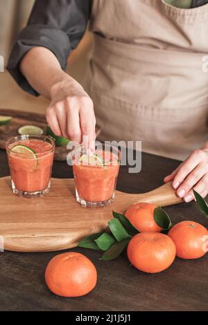 Nahaufnahme einer nicht erkennbaren Frau auf der Schürze, die an der Küchentheke steht und den frischen Mandarinensaft mit Limettenscheiben dekoriert Stockfoto