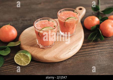 Nahaufnahme eines Holzbretts mit Gläsern frischen Grapefruitsafts, dekoriert mit Limettenscheibe, Mandarinen und Blättern auf einem Holztisch Stockfoto