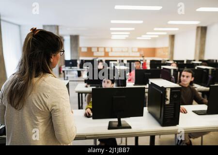 Reife Lehrerin, die mit Schülern im Computerraum der Schule arbeitet - Fokus auf die Schulter der Frau Stockfoto