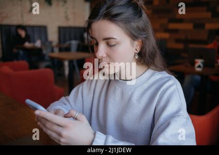 Glücklich lächelnd wunderschöne Frau in modischen Brillen lesen angenehme Textnachricht auf dem Handy, während im Café während der Freizeit sitzen. Stockfoto