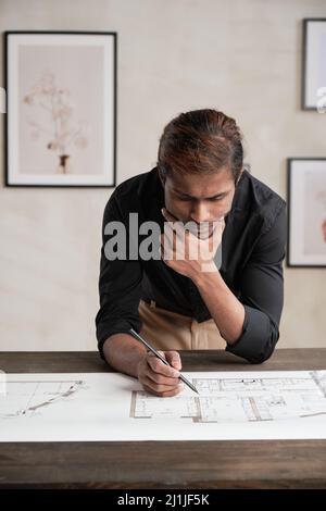 Fokussierter junger indischer Gebäudedesigner in schwarzem Hemd, der sich auf einen Holztisch lehnte und das Kinn in der Hand hielt, während er den Bauplan untersuchte Stockfoto
